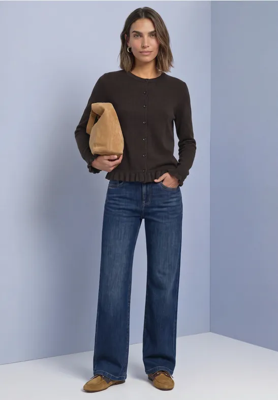 Frau trägt braunen Pullover, weite Jeans und hält eine braune Tasche vor schlichtem hellblauem Hintergrund.