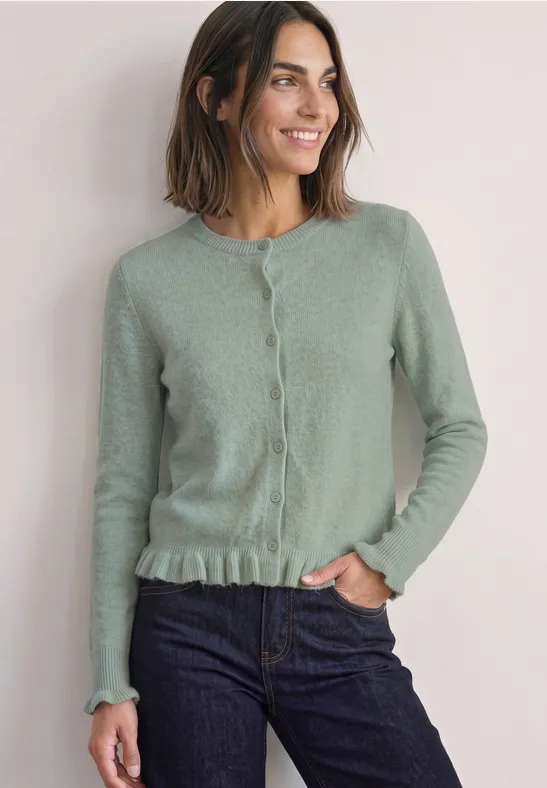 Frau mit schulterlangem Haar trägt grüne Strickjacke mit Knopfleiste und Rüschenbund, lächelt leicht.