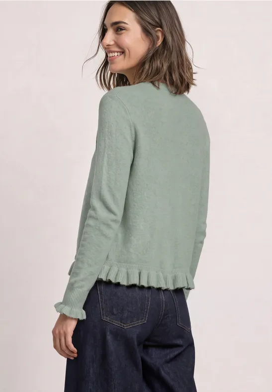 Eine Frau mit schulterlangem Haar, die über die Schulter lächelnd eine hellgrüne Strickjacke trägt.