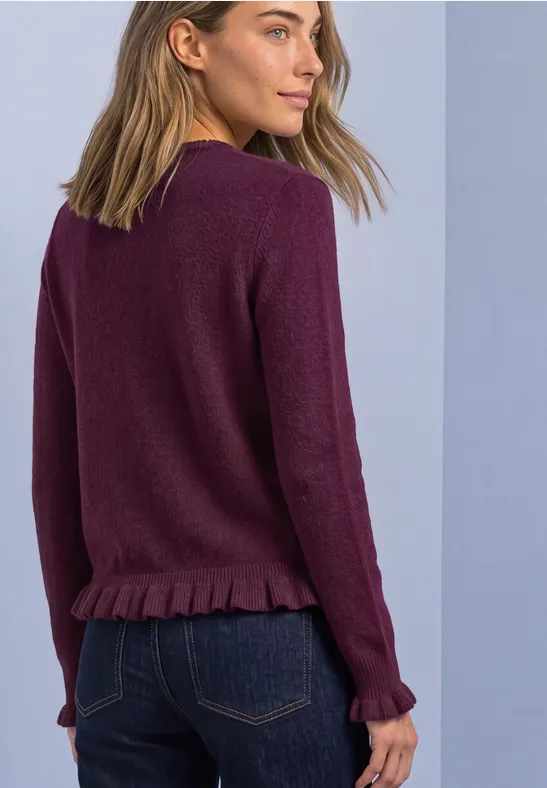 Frau in seitlicher Rückenansicht trägt violetten Pullover mit Rüschensaum und dunkelblaue Jeans vor hellem Hintergrund.