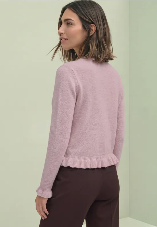 Eine Frau in rosa Strickpullover und dunkler Hose, im Profil vor hellem Hintergrund.