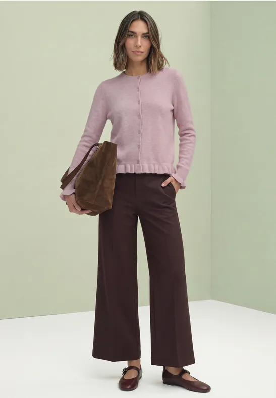 Frau trägt rosafarbenen Strickcardigan mit Rüschen, weite dunkle Hose, braune Mules, hält eine braune Tasche.