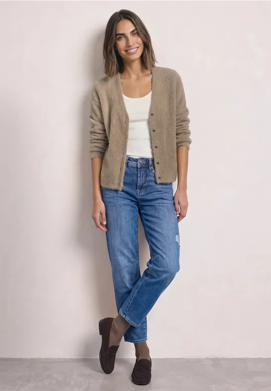 Frau in Jeans, weißem Shirt und beiger Strickjacke steht entspannt vor hellem Hintergrund.
