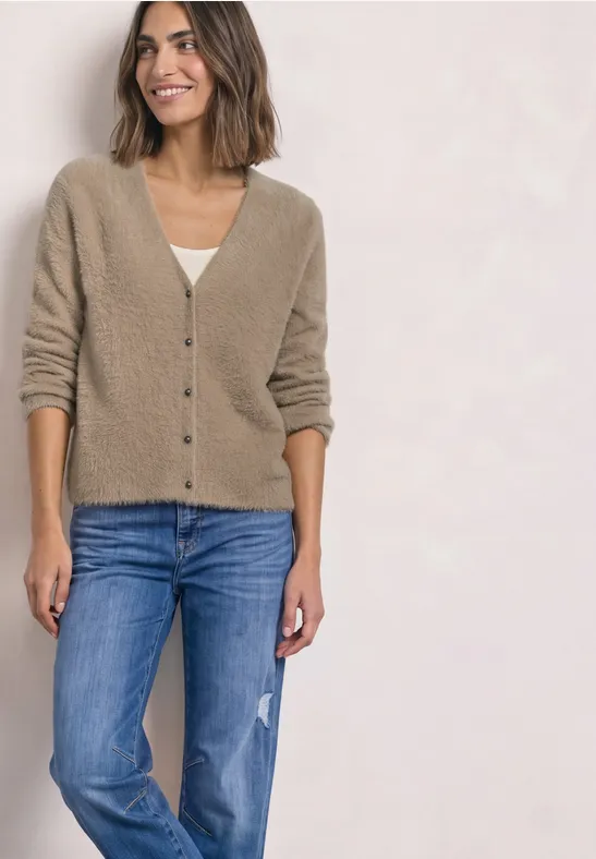 Eine Frau trägt eine beige Strickjacke und blaue Jeans, steht lächelnd vor hellem Hintergrund.
