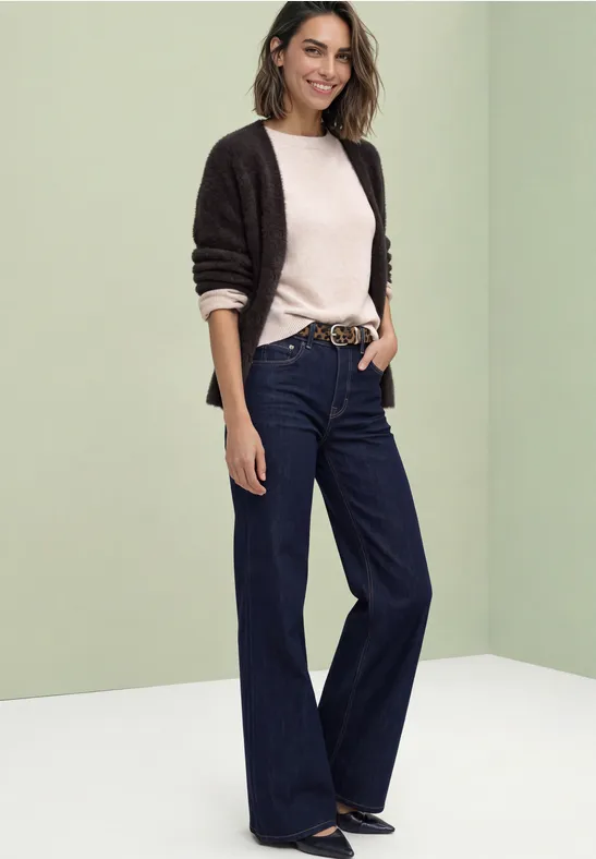 Eine Frau in weitem dunkelblauen Jeans, hellem Pullover und schwarzer Strickjacke vor grünlich-weißer Kulisse.