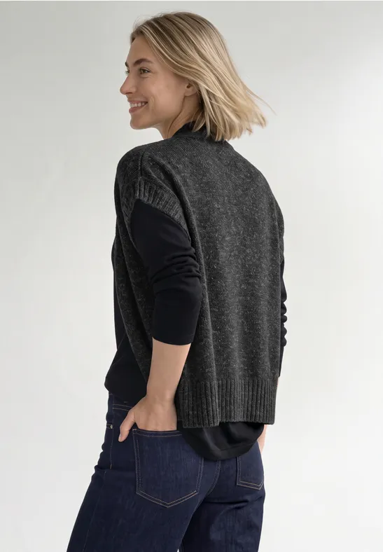 Eine Frau in dunklen Jeans, schwarzem Langarmshirt und grauer Strickweste geht seitlich, lächelnd.