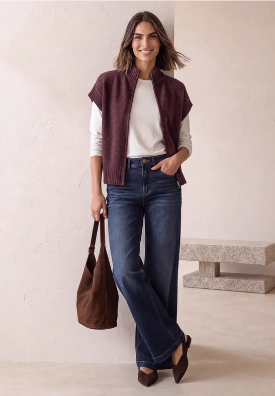 Frau in dunklen Jeans, weißem Shirt, weinrotem Pullover um die Schultern, mit brauner Tasche.