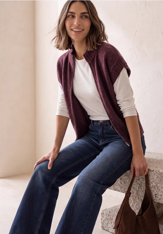 Lächelnde Frau in weißem Shirt und dunkelblauen Jeans, mit bordeauxfarbener Jacke über den Schultern und passender Tasche.