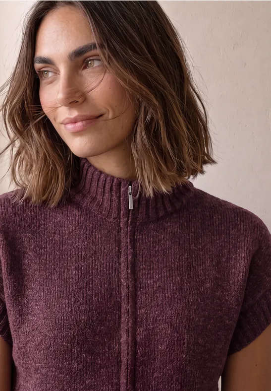 Nahaufnahme einer Frau mit schulterlangen braunen Haaren und einem kurzärmligen, violetten Strickpullover mit Reißverschluss.