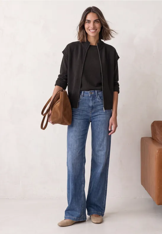 Frau in schwarzer Jacke, schwarzem Top und blauen Jeans hält eine braune Tasche vor hellem Hintergrund.