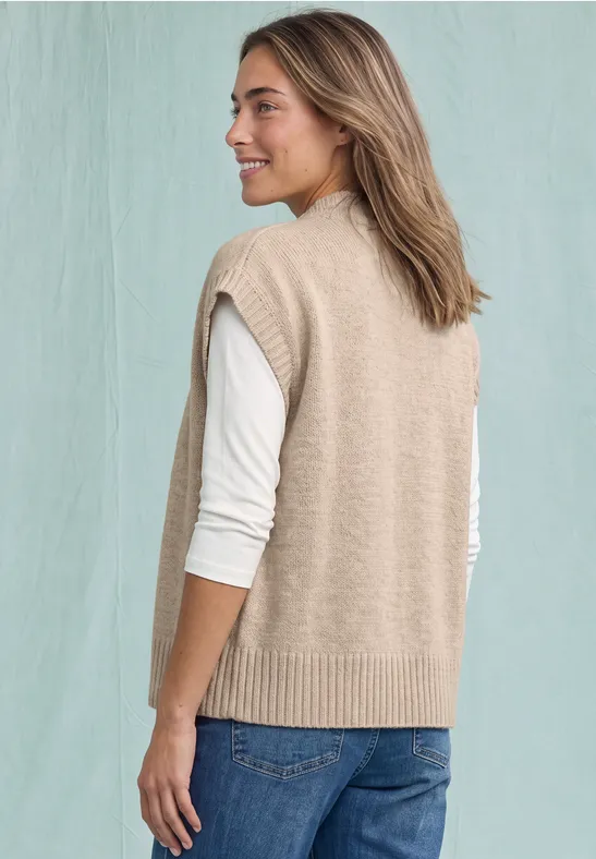 Een vrouw draagt een beige gebreid vest over een wit longsleeve shirt en jeans, glimlachend opzij kijkend.