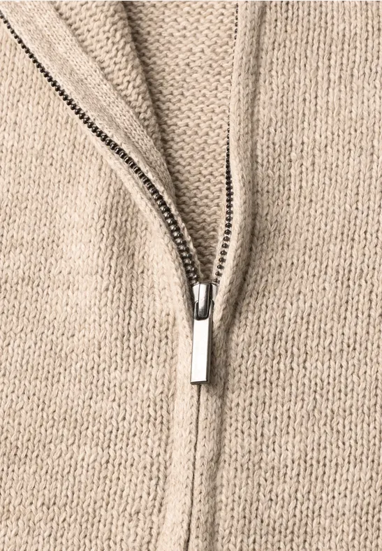 Close-up van een beige gebreide trui met een deels open zilverkleurige ritssluiting