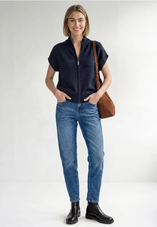 Junge Frau in blauer Jeans, schwarzem Kurzarm-Oberteil und Stiefeln, mit brauner Schultertasche vor weißem Hintergrund.