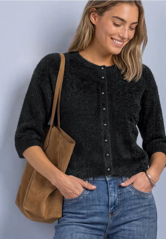 Frau trägt schwarzen flauschigen Cardigan, blaue Jeans und braune Schultertasche, lächelt vor hellem Hintergrund.