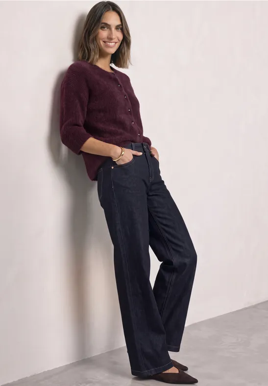 Eine lächelnde Frau lehnt an einer Wand und trägt einen weinroten Pullover sowie weite, dunkle Hosen.