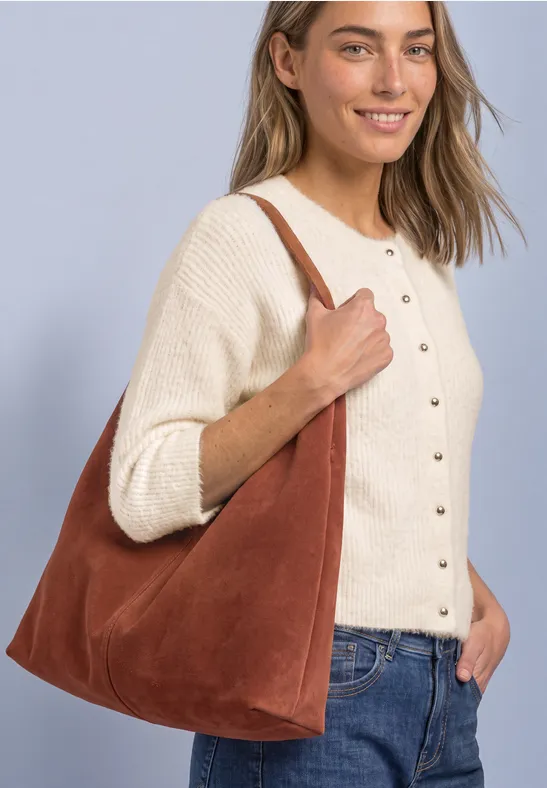 Eine Frau trägt lächelnd eine braune Wildleder-Hobo-Tasche an der Schulter, dazu cremefarbener Cardigan und Jeans.