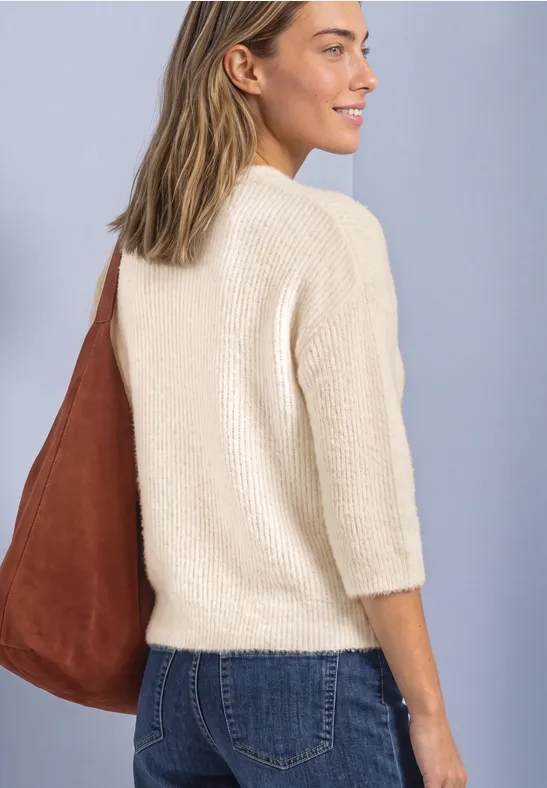 Frau in weißem Pullover und Jeans mit brauner Schultertasche blickt seitlich.