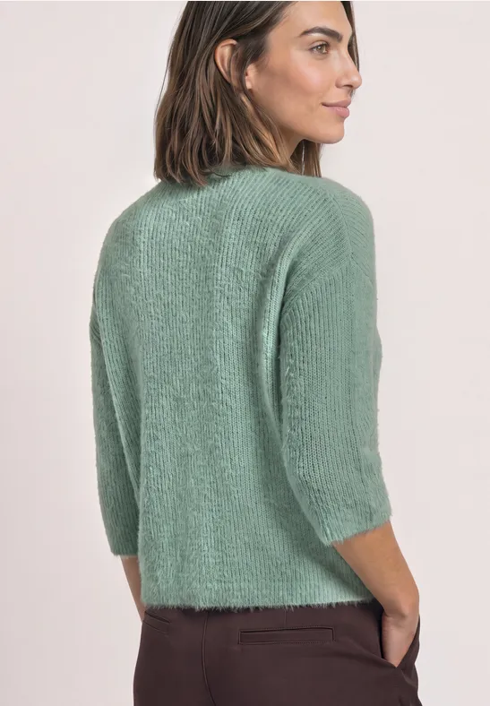 Frau in flauschigem mintgrünem 3/4-Pullover und dunkelroten Hosen, seitlich abgewandt.