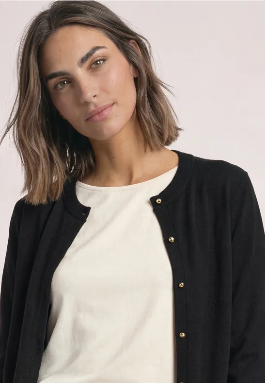 Frau mit braunen Haaren trägt schwarzen Cardigan und weißen Pullover, lächelnd.