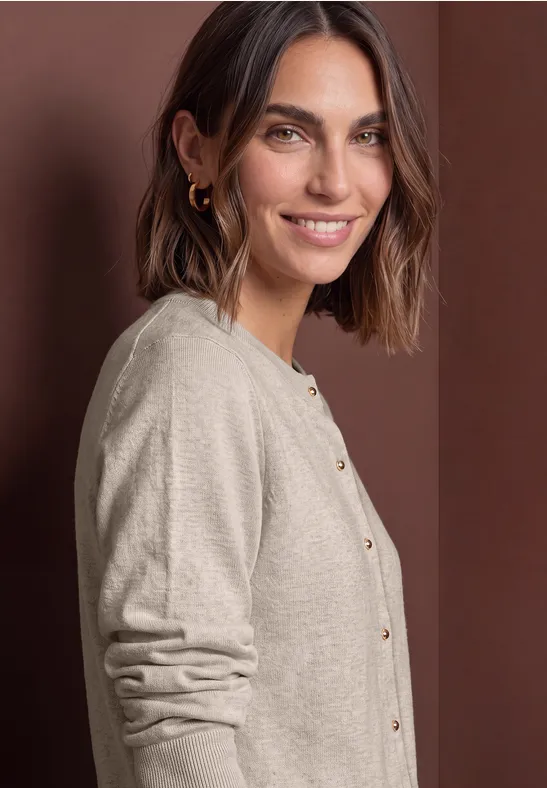 Kurzhaarige, lächelnde Frau mit braunen Haaren, Ohrringen und beige Strickjacke mit goldenen Knöpfen vor brauner Wand.