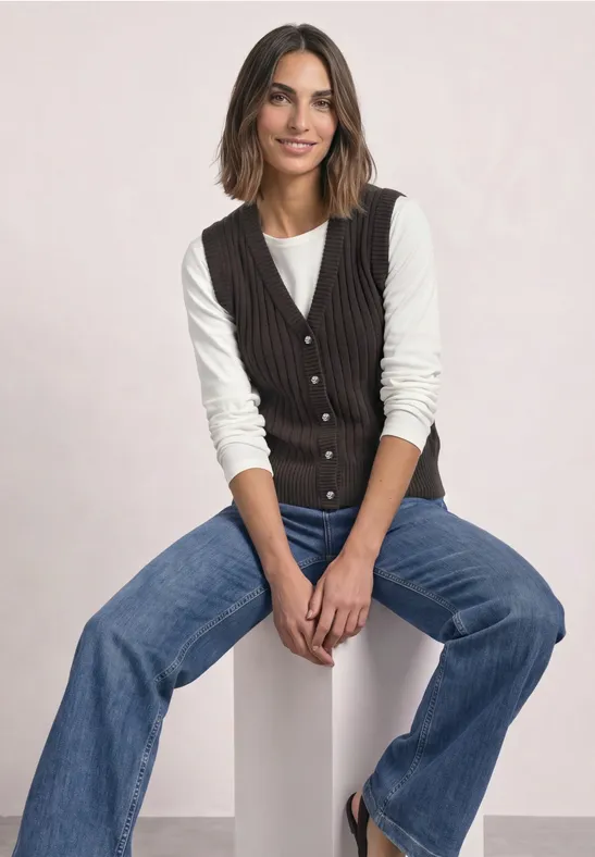 Eine lächelnde Frau sitzt auf einem weißen Block, trägt Jeans, weißen Pullover und dunkle Weste.