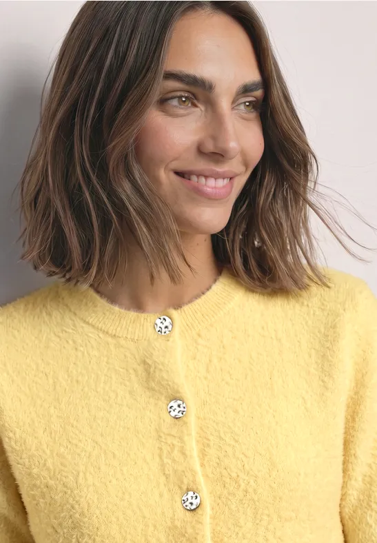 Eine lächelnde Frau mit mittellangem braunem Haar trägt einen gelben Strickcardigan mit verspielten Knöpfen.
