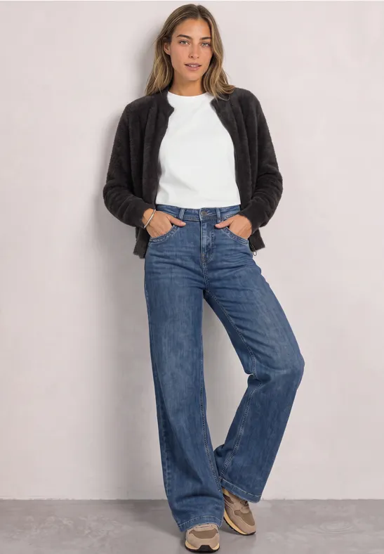Frau in weißem T-Shirt, schwarzer Jacke und weitem Jeans lehnt lässig vor einer hellen Wand.