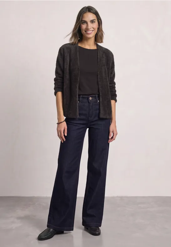 Frau in dunkler Jacke, schwarzem Shirt und weitem Jeans, lächelnd vor hellem Hintergrund.