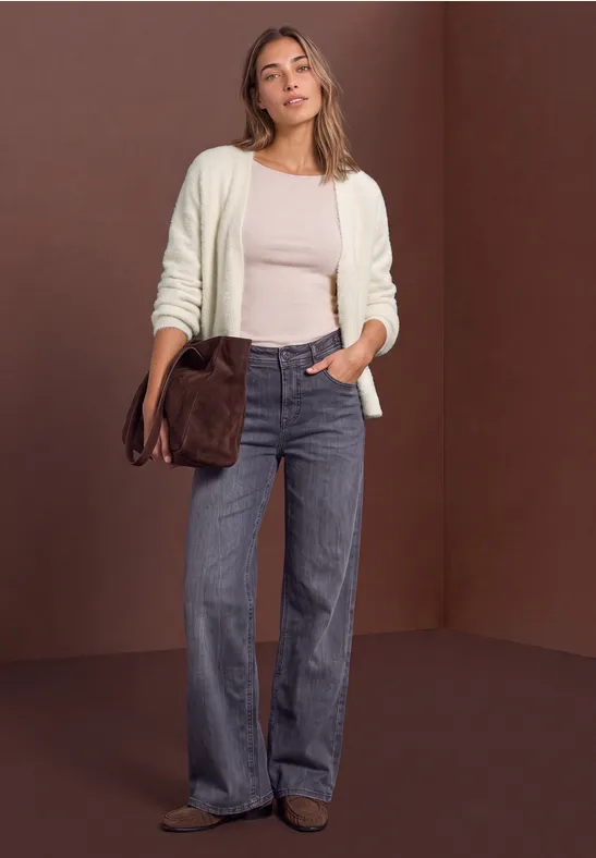 Frau mit weitem Jeans, beigem Top und weißer Strickjacke, hält eine braune Tasche vor braunem Hintergrund.