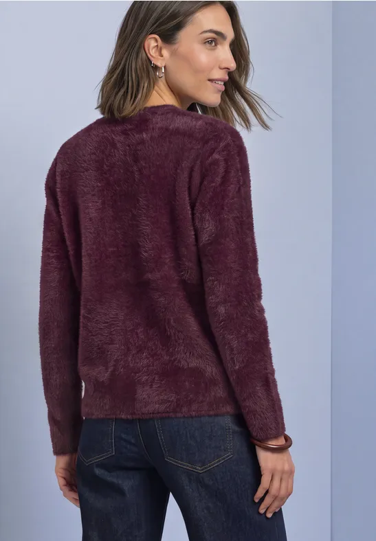 Frau von hinten in dunkelblauer Jeans und beerenfarbenem, flauschigem Pullover vor neutralem Hintergrund.