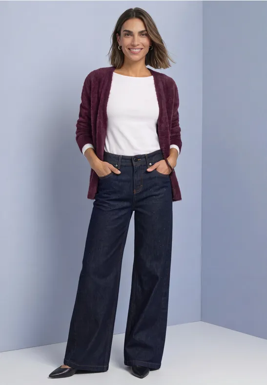 Eine Frau lächelnd in weißem Top, burgunderfarbener Strickjacke und weitem Jeans vor hellem Hintergrund.