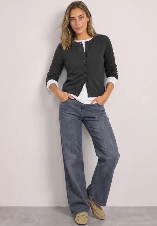Frau in schwarzem Cardigan, weißem Shirt und weiten Jeans lehnt lässig vor einer grauen Wand.
