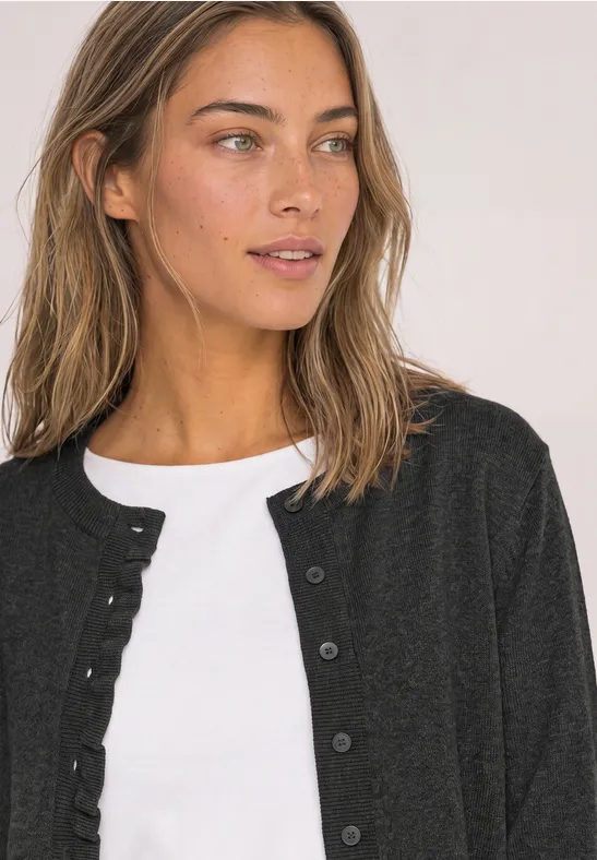 Frau mit braunen Haaren trägt weißen Top und schwarzen Cardigan, blickt seitlich.