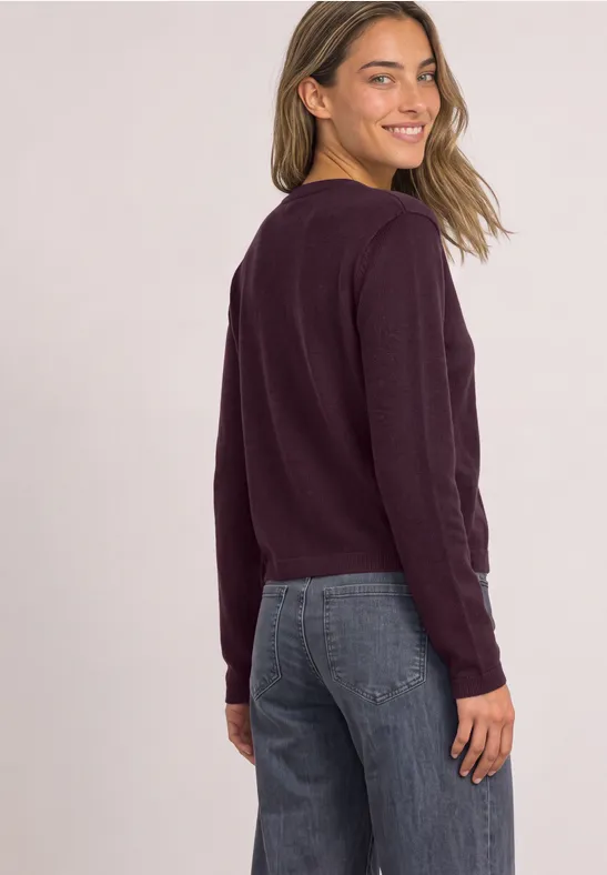 Eine Frau blickt über die Schulter und lächelt, trägt einen kastanienbraunen Pullover und blaue Jeans.