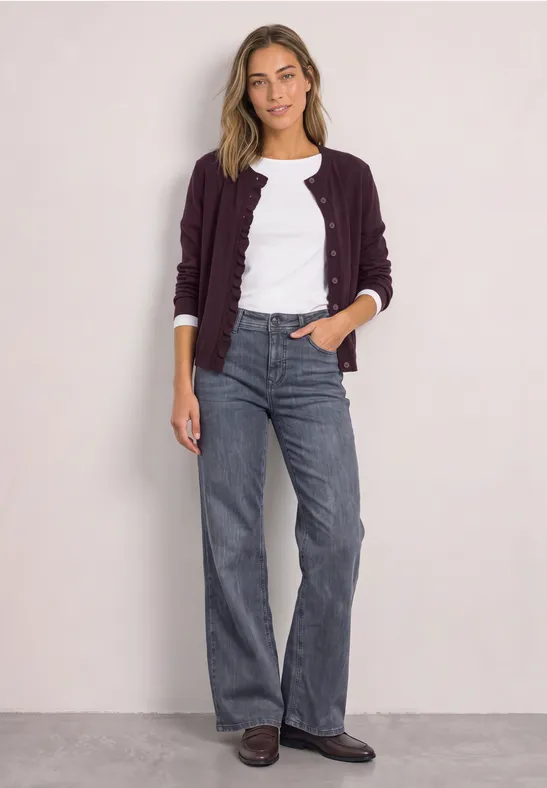 Eine Frau steht vor heller Wand, trägt weiße Bluse, bordeauxfarbene Strickjacke, weite graue Jeans und braune Schuhe.