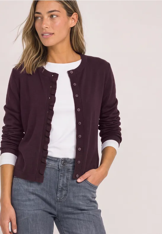 Frau in lila Cardigan mit Rüschen am Saum, weißem Shirt und grauer Hose.