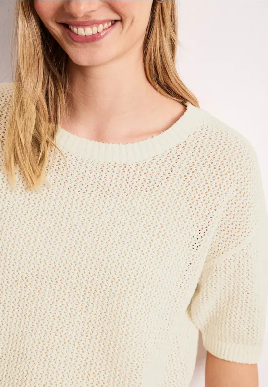Close-up van een glimlachende vrouw in een crème-kleurige, losgebreide trui met korte mouwen.