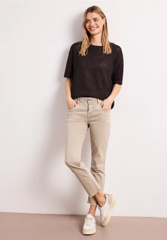 Femme en haut noir, pantalon beige et baskets blanches, debout contre un mur blanc, mains dans les poches.