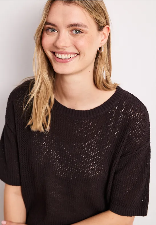 Femme portant un pull en maille noire ample, motif transparent, cheveux blonds, fond clair.