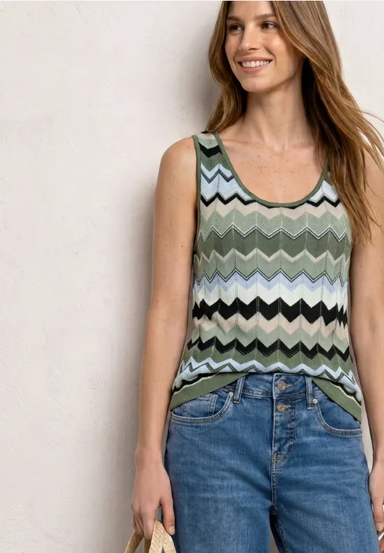 Frau trägt ärmelloses, grün-schwarz-weißes Zickzack-Oberteil und blaue Jeans, hält ein Handy.