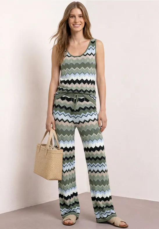 Frau in ärmellosem Jumpsuit mit pastellgrünem Zickzackmuster und Strohtasche.