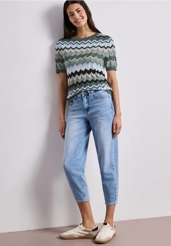 Vrouw in zigzagtrui, lichtblauwe jeans en instappers voor een neutrale achtergrond.