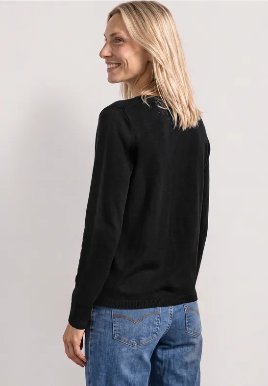Blonde Frau trägt schwarzen Pullover und blaue Jeans, steht seitlich vor neutralem Hintergrund.