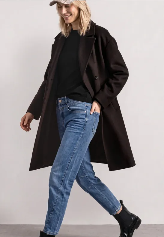Frau in lässigem Outfit mit langem schwarzen Mantel, schwarzem Pullover, blauen Jeans, schwarzen Boots und weißer Mütze.