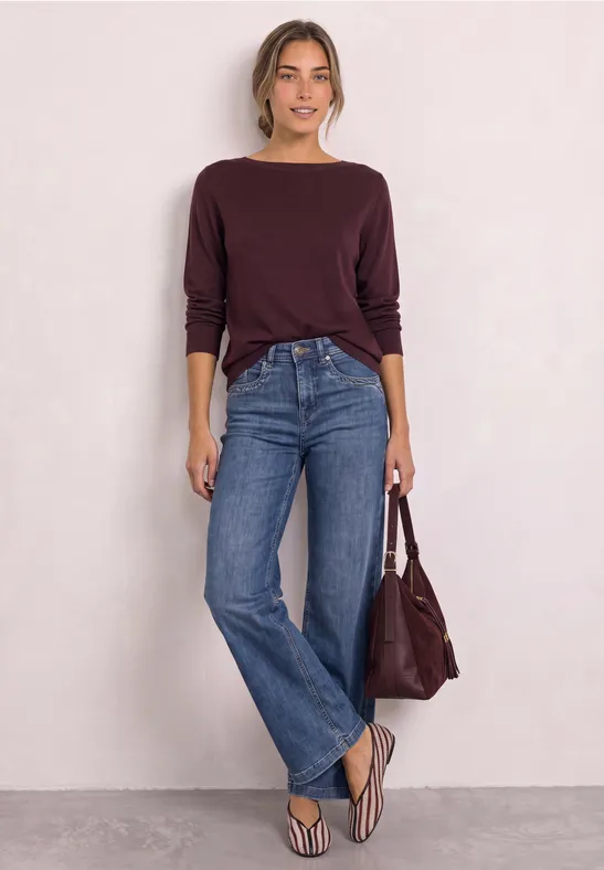 Vrouw in bordeaux trui, blauwe wijde jeans, slippers, bijpassende tas, staand tegen een witte muur.