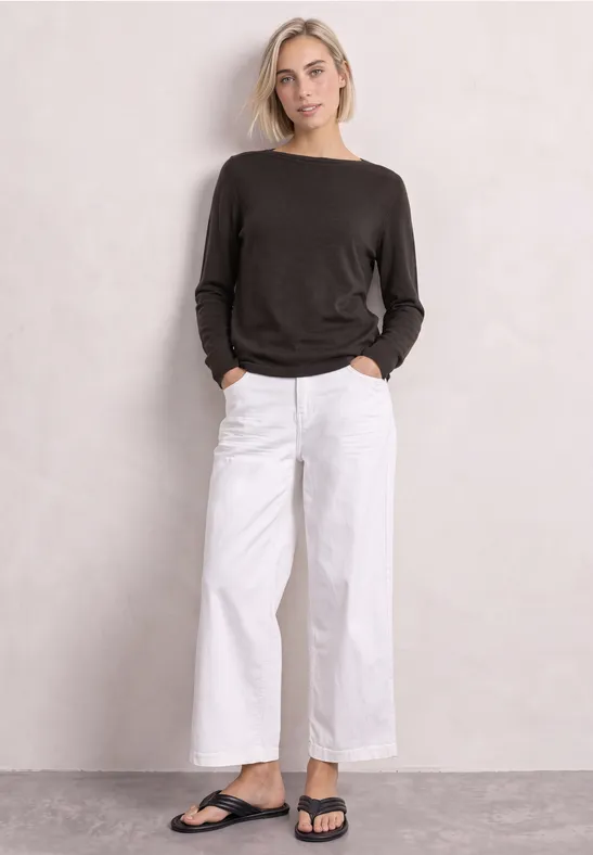 Frau in schwarzem Pullover, weißer weiter Hose und schwarzen Sandalen vor neutralem Hintergrund.