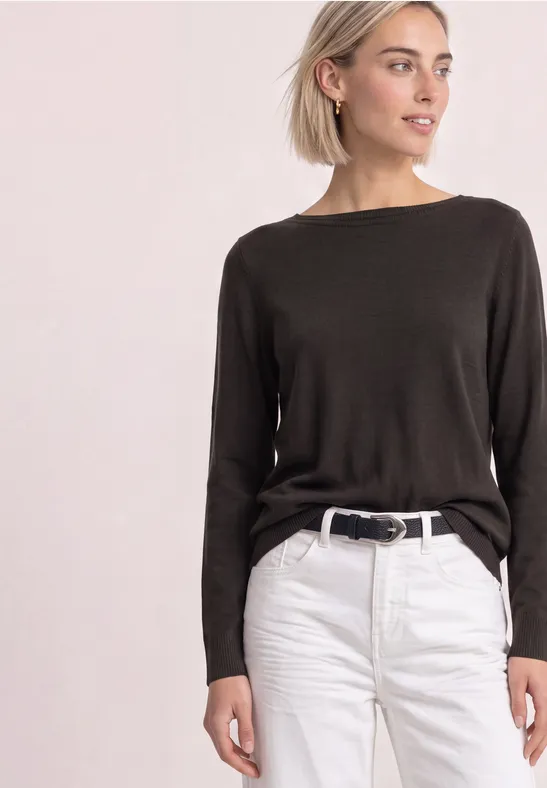 Frau in kastanienbraunem Pullover mit langen Ärmeln und weißen Jeans, schwarzem Gürtel vor hellem Hintergrund.