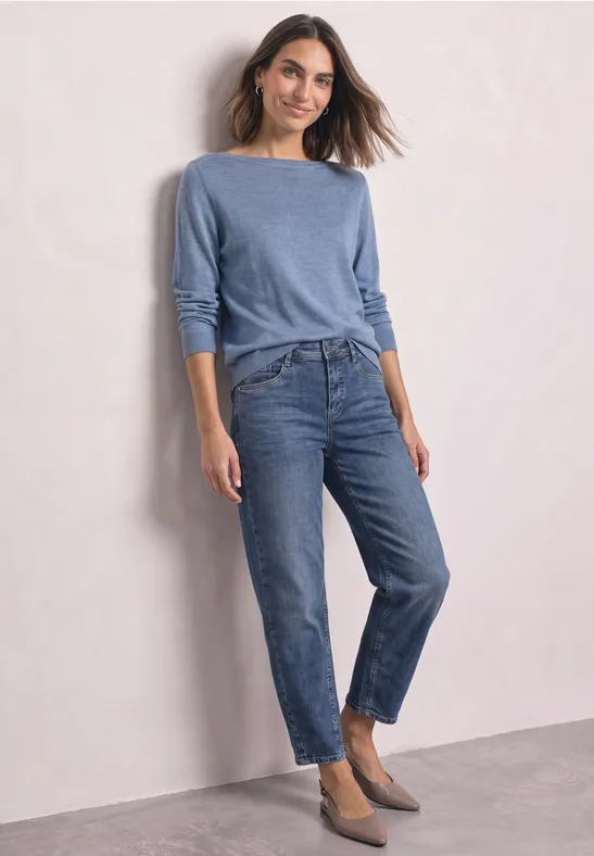 Frau mit blauem Pullover und Jeans, steht vor heller Wand.
