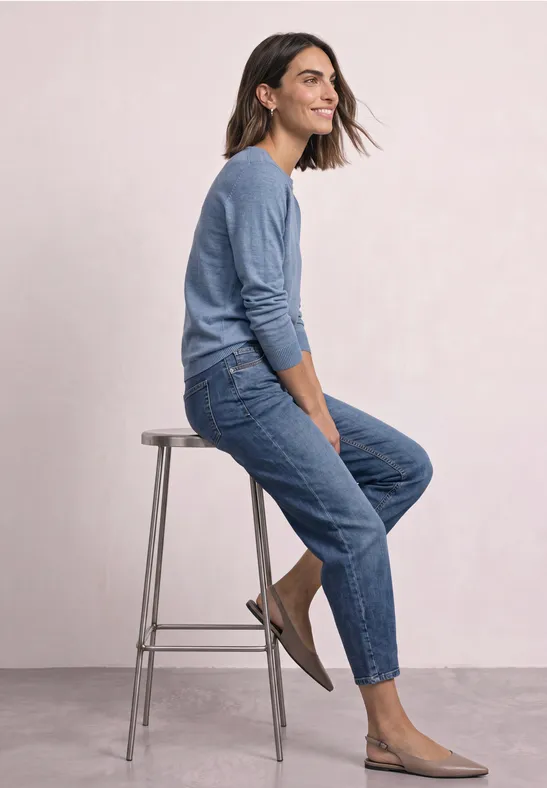 Frau sitzt seitlich auf einem Hocker, trägt blauen Pullover, Jeans und flache Schuhe vor hellem Hintergrund.