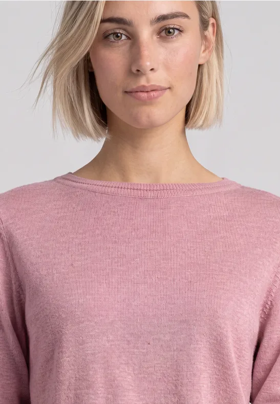 Frau mit kurzen blonden Haaren trägt einen rosafarbenen Pullover vor neutralem Hintergrund.
